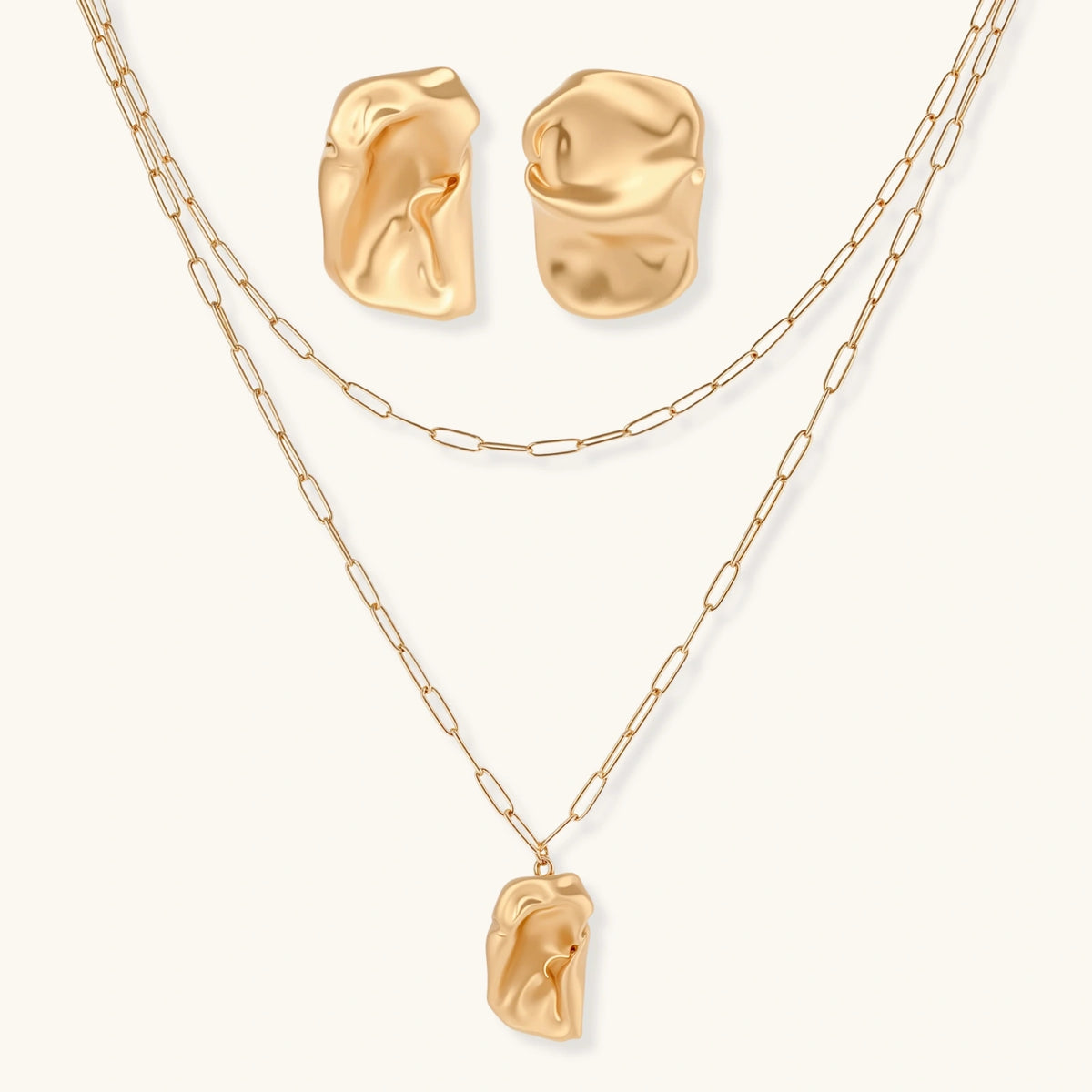 Sculptural gold-toned jewelry set with organic fluid shapes including earrings and layered chain necklace on cream background.